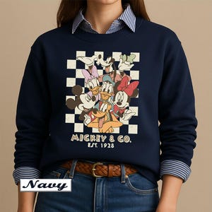 May include: A navy blue crewneck sweatshirt featuring a graphic print of Mickey Mouse, Minnie Mouse, Donald Duck, Daisy Duck, Goofy, and Pluto over a white and navy checkered background with the text MICKEY & CO. EST. 1928 printed below the characters.