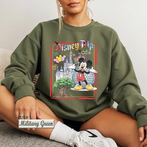 May include: A military green long-sleeve sweatshirt featuring a graphic print of Mickey Mouse, a castle, a tree, and the text Disney Trip 2024. The design includes colorful fireworks and a geometric sphere, all framed within a red rectangular border.