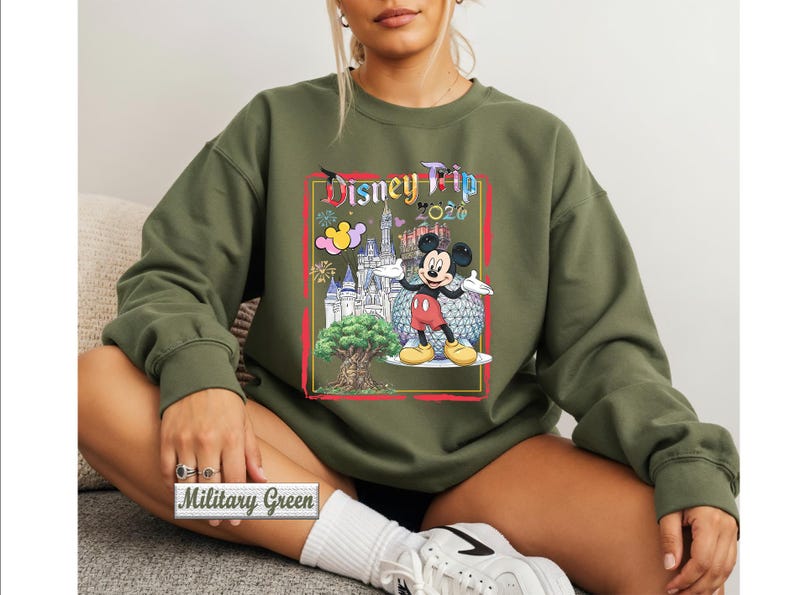 May include: A military green long-sleeve sweatshirt featuring a graphic print of Mickey Mouse, a castle, a tree, and the text Disney Trip 2024. The design includes colorful fireworks and a geometric sphere, all framed within a red rectangular border.