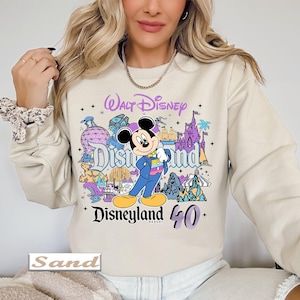 Op de afbeelding: Een zandkleurige sweater met ronde hals en een kleurrijke grafische print van Mickey Mouse in een blauw pak, omringd door Disneyland Resort-iconen en de tekst Walt Disney Disneyland 40. Het ontwerp bevat paarse, blauwe en gele accenten.
