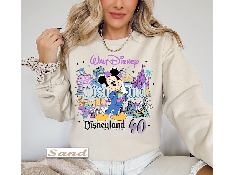 Op de afbeelding: Een zandkleurige sweater met ronde hals en een kleurrijke grafische print van Mickey Mouse in een blauw pak, omringd door Disneyland Resort-iconen en de tekst Walt Disney Disneyland 40. Het ontwerp bevat paarse, blauwe en gele accenten.