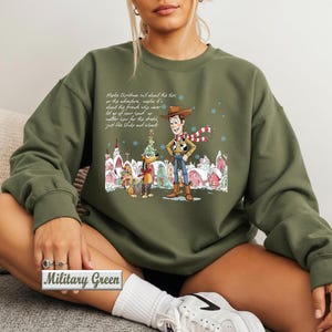 May include: A military green crewneck sweatshirt featuring a graphic print of Woody and Slinky Dog from Toy Story in a snowy village setting. The design includes a holiday theme with text above the characters and a small label reading Military Green.