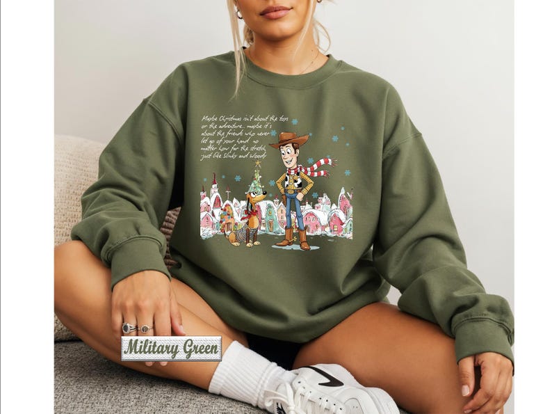 May include: A military green crewneck sweatshirt featuring a graphic print of Woody and Slinky Dog from Toy Story in a snowy village setting. The design includes a holiday theme with text above the characters and a small label reading Military Green.