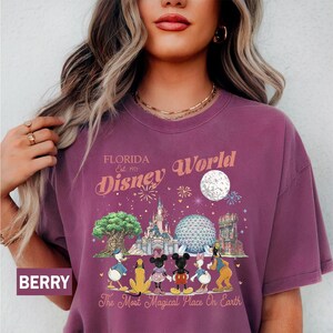 May include: A berry-colored crew neck t-shirt featuring a graphic print of Disney World landmarks and characters. The text reads Florida Est. 1971 Disney World and The Most Magical Place On Earth, accompanied by illustrations of the castle and park icons.