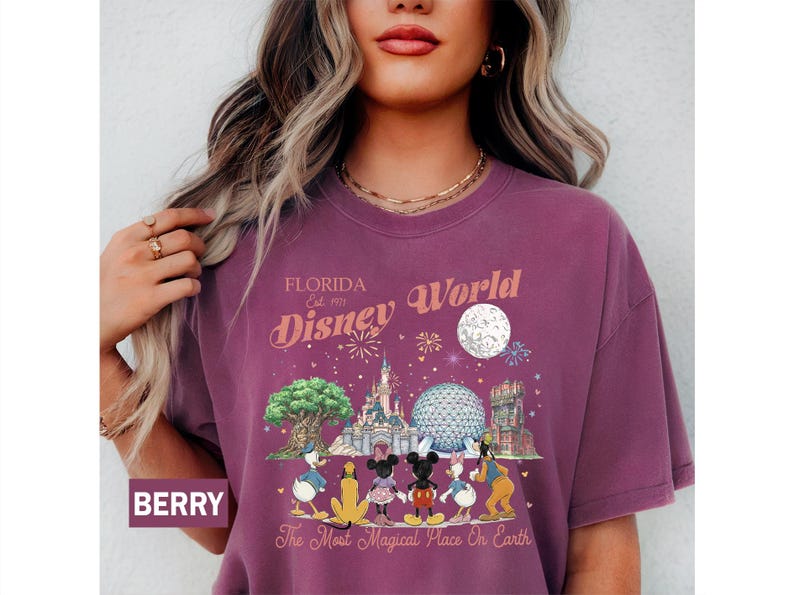 May include: A berry-colored crew neck t-shirt featuring a graphic print of Disney World landmarks and characters. The text reads Florida Est. 1971 Disney World and The Most Magical Place On Earth, accompanied by illustrations of the castle and park icons.