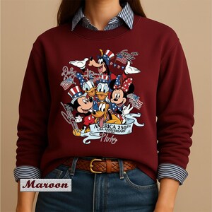 May include: A maroon long-sleeve sweatshirt featuring a graphic print of Disney characters including Mickey Mouse, Minnie Mouse, Donald Duck, Daisy Duck, Goofy, and Pluto wearing patriotic hats. The text reads AMERICA 250TH USA ANNIVERSARY 1776-2026.