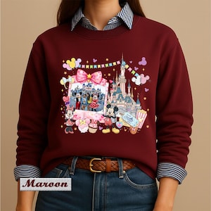 May include: A maroon long-sleeve sweatshirt featuring a graphic print of a castle, a photo frame with cartoon characters, and various colorful icons including balloons, a cupcake, and a ticket. The sweatshirt has a crew neckline and ribbed cuffs.