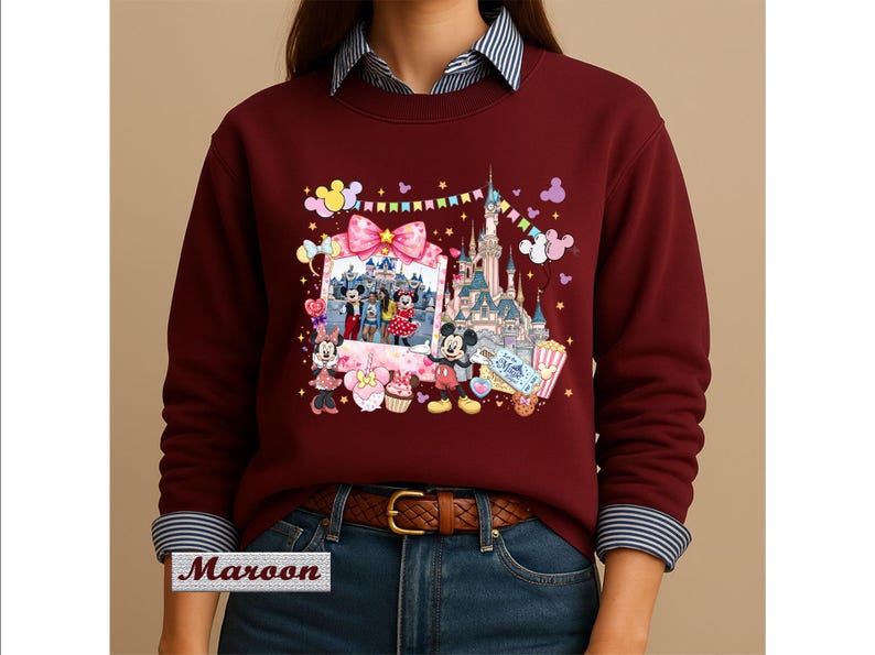 May include: A maroon long-sleeve sweatshirt featuring a graphic print of a castle, a photo frame with cartoon characters, and various colorful icons including balloons, a cupcake, and a ticket. The sweatshirt has a crew neckline and ribbed cuffs.