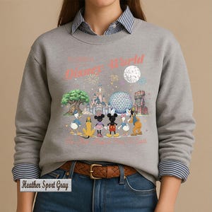 May include: A heather sport gray crewneck sweatshirt featuring a graphic of Disney characters including Mickey Mouse, Minnie Mouse, Donald Duck, Daisy Duck, Pluto, and Goofy standing before park landmarks with the text Disney World and The Most Magical Place On Earth.