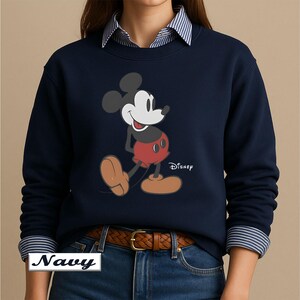May include: A navy blue long-sleeve sweatshirt featuring a graphic of Mickey Mouse with a black head, red shorts, and tan shoes. The word Disney is printed in white next to the character. The sweatshirt is worn over a blue and white striped shirt.