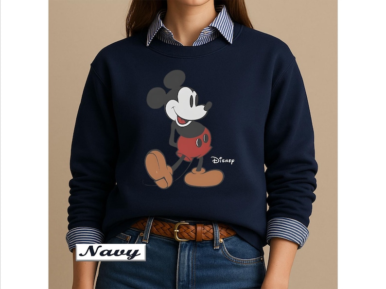 May include: A navy blue long-sleeve sweatshirt featuring a graphic of Mickey Mouse with a black head, red shorts, and tan shoes. The word Disney is printed in white next to the character. The sweatshirt is worn over a blue and white striped shirt.