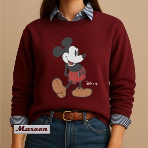 May include: A maroon long-sleeve sweatshirt featuring a graphic of Mickey Mouse with the Disney logo. The sweatshirt is worn over a blue and white striped collared shirt, paired with blue jeans and a brown leather belt with a gold buckle.