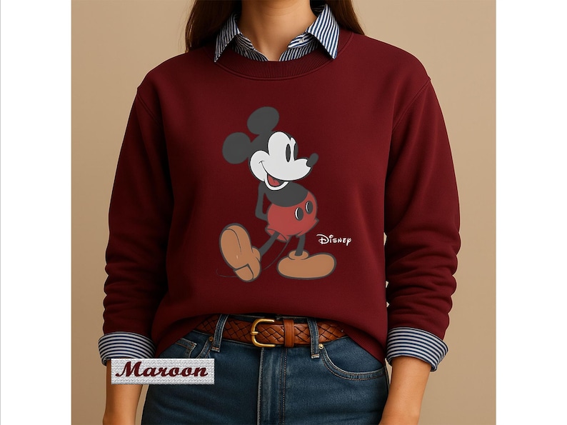 May include: A maroon long-sleeve sweatshirt featuring a graphic of Mickey Mouse with the Disney logo. The sweatshirt is worn over a blue and white striped collared shirt, paired with blue jeans and a brown leather belt with a gold buckle.