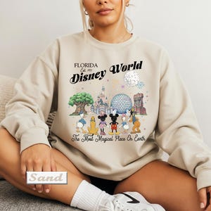 May include: A sand-colored crewneck sweatshirt featuring a graphic print of Disney World landmarks and characters. The text reads Florida Est. 1971 Disney World and The Most Magical Place On Earth. The design includes Mickey, Minnie, and friends.