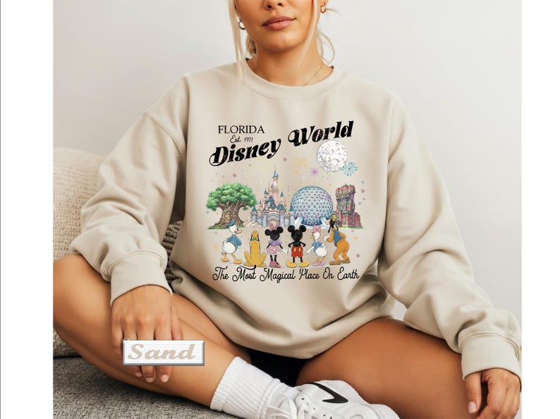 May include: A sand-colored crewneck sweatshirt featuring a graphic print of Disney World landmarks and characters. The text reads Florida Est. 1971 Disney World and The Most Magical Place On Earth. The design includes Mickey, Minnie, and friends.