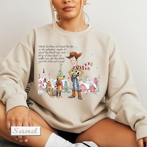 May include: A beige crewneck sweatshirt featuring a graphic print of Woody and Slinky Dog from Toy Story in a winter village setting. The text reads: Maybe Christmas isn't about the toys or the adventure, maybe it's about the friends who never let go of your hand no matter how far the stretch, just like Slinky and Woody.