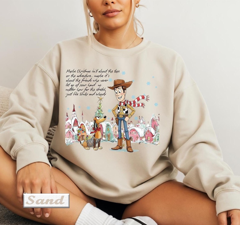 May include: A beige crewneck sweatshirt featuring a graphic print of Woody and Slinky Dog from Toy Story in a winter village setting. The text reads: Maybe Christmas isn't about the toys or the adventure, maybe it's about the friends who never let go of your hand no matter how far the stretch, just like Slinky and Woody.