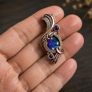 Puede incluir: Un colgante de alambre trenzado con una piedra central redonda facetada en azul y morado iridiscente, acentuado por dos pequeños cabujones morados. El marco de metal está hecho de alambre de cobre con bucles y patrones intrincados.
