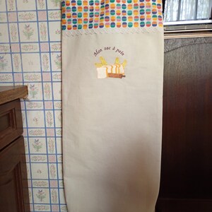 Peut inclure: Un sac à pain beige avec un motif à pois colorés en haut, orné d'une bordure en dentelle blanche. Le sac porte la mention brodée "Mon sac à pain" ainsi que des images de blé, de pain et d'un rouleau à pâtisserie. Il a une anse pour suspendre.