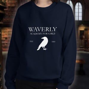以下が含まれることがあります： ネイビーブルーの長袖スウェットシャツ。胸元には「WAVERLY ACADEMY FOR GIRLS」と「EST. 1866」という白い文字がプリントされており、その中央には横向きのワタリガラスの白いシルエットが描かれています。