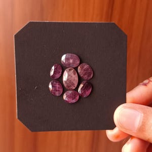 May include: A selection of oval-cut, purplish-red gemstones displayed on a black octagonal card. The gemstones have slight variations in colour and clarity, with some being more translucent. The arrangement is held by a hand.