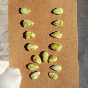 May include: A collection of sixteen pale green, teardrop-shaped gemstones with brown inclusions, arranged on a brown card. The stones vary slightly in size and color, with some showing more brown marbling than others. The gemstones are displayed in two columns.