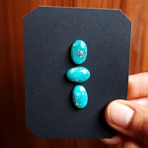 May include: Three oval-shaped turquoise gemstones with a vibrant blue-green hue and natural matrix patterns. The stones are displayed on a black background, showcasing their color and texture. The gemstones are likely for jewelry making.