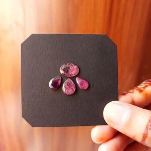 Op de afbeelding: Vier roze edelstenen van verschillende vormen worden gepresenteerd op een zwart oppervlak. De edelstenen omvatten een ovaal, een gefacetteerde peervorm en twee druppelvormen. De edelstenen worden vastgehouden door een hand met henna-tatoeages.