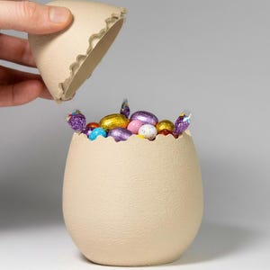Könnte beinhalten: Ein beigefarbener, eiförmiger Behälter mit strukturierter Oberfläche, der teilweise geöffnet ist, um bunte, verpackte Süßigkeiten zu zeigen. Der Deckel hat einen gezackten Rand. Der Behälter ist mit folienverpackten Schokoladeneiern gefüllt.