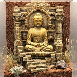 Könnte beinhalten: Eine goldene Buddha-Statue in meditativer Haltung sitzt in einer detaillierten steinartigen Tempelstruktur. Die Szene umfasst einen rötlich-braunen, strukturierten Untergrund, kleine grüne Sukkulenten, getrocknete Gräser und dunkelgraue Steine in einem Glasgehäuse.