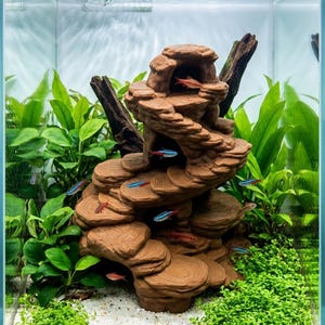 May include: An aquarium scene featuring a brown, rock-like structure with multiple levels, surrounded by green plants and small, colorful fish. The tank has a white gravel base and is filled with clear water.