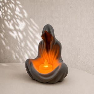 Pode incluir: Uma escultura de cerâmica cinza-escura de uma figura encapuzada em uma pose sentada e meditativa. A figura apresenta um centro oco que contém uma pequena vela de chá LED laranja brilhante, emitindo uma luz quente de dentro do manto.