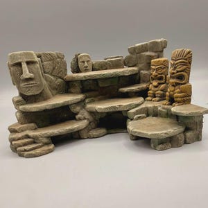 Könnte beinhalten: Ein mehrstöckiges, steinfarbenes Aquarium-Ornament mit einem großen Moai-Statuenkopf auf der linken Seite, zwei kleineren geschnitzten Tiki-Figuren auf der rechten Seite und einem zentralen menschlichen Gesichtsrelief, integriert in eine gestufte Struktur.
