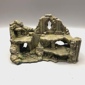 Può includere: Un ornamento decorativo per acquario che raffigura una struttura in rovina di pietra a più livelli con texture invecchiate, archi e colonne. L'oggetto è colorato in tonalità tenui di verde, grigio e marrone, con camere cave e piattaforme.