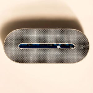 K&ouml;nnte beinhalten: Ein ovales Objekt mit gemusterter Oberfl&auml;che und rechteckiger &Ouml;ffnung. Das Objekt hat einen beigen Rand und ein schwarz-wei&szlig;es geometrisches Muster. Die &Ouml;ffnung zeigt einen blauen Innenraum. Der Hintergrund ist beige.