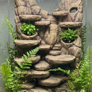 Może przedstawiać: Pionowe tło do terrarium z teksturowaną, jasnobrązową strukturą przypominającą skałę, z wieloma zintegrowanymi okrągłymi platformami i kieszeniami na rośliny. Jasnozielone paprocie i małe rośliny liściaste są umieszczone wewnątrz struktury.
