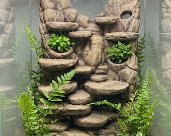 May include: A vertical terrarium background featuring a textured, light brown rock-like structure with multiple integrated circular platforms and recessed planting pockets. Bright green ferns and small leafy plants are nestled within the structure.