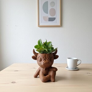 Pode incluir: Um vaso de plantas marrom em forma de vaca Highland com plantas verdes, sentado em uma mesa de madeira clara. Uma xícara de chá e um pires brancos estão à direita. Uma impressão de arte abstrata emoldurada está pendurada na parede branca ao fundo.