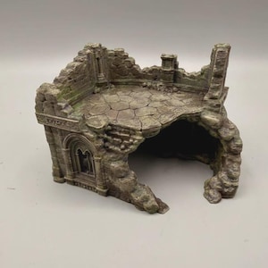 Pode incluir: Uma estrutura de ruína de pedra em miniatura com um acabamento texturizado em cinza e verde desgastado. A peça inclui um piso circular pavimentado, seções de parede em ruínas com detalhes arquitetônicos e uma grande abertura em arco na base.