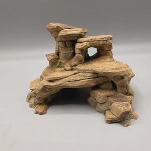 Op de afbeelding: Een beige en bruin rotsformatie aquariumornament met een structuur van meerdere niveaus, een holle grotopening aan de basis, getextureerde oppervlakken die lijken op gelaagde steen en een kleine boogvormige opening aan de rechterbovenkant.