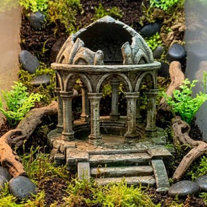 Könnte beinhalten: Ein grauer, verwitterter, runder Steingartenpavillon mit bogenförmigen Säulen und einem Kuppeldach, der über eine Treppe am Eingang verfügt und in eine Miniaturlandschaft aus dunkler Erde, grünem Moos, kleinen Steinen und Treibholz eingebettet ist.
