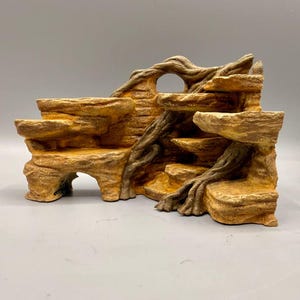 May include: A tan and brown terrarium ornament featuring a multi-level rock formation with flat ledges and integrated tree root textures. The structure includes a small circular opening near the top and a hollow archway at the base for shelter.