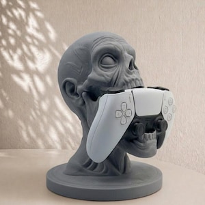 Pode incluir: Um suporte cinzento em forma de caveira para comando de jogos. O suporte foi concebido para acomodar um comando de jogos branco e preto. A caveira tem detalhes e assenta numa base circular. O comando é segurado na boca da caveira.