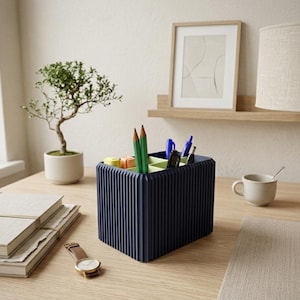 Pode incluir: Um organizador de secretária azul marinho com linhas verticais, contendo lápis, canetas e artigos de papelaria. Um pequeno bonsai, livros e um relógio de pulso estão na secretária de madeira clara. Uma imagem emoldurada e uma lâmpada estão no fundo.