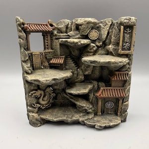 Könnte beinhalten: Ein dekorativer steinfarbener Präsentationsständer mit einer skulptierten Berglandschaft, abgestuften Plattformen, Miniatur-Pagodenbauten mit rotbraunen Dächern, einem Drachenrelief und vertikalen rechteckigen Tafeln mit eingraviertem Text.