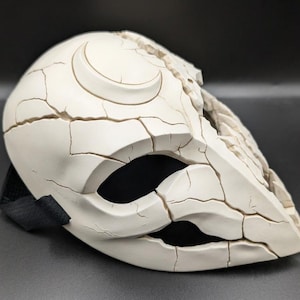 Op de afbeelding: Een crèmekleurig masker met een gebarsten, verweerde textuur en een verhoogd maansikkel-symbool op het voorhoofd. Het masker heeft grote, donkere oogopeningen en een grillig, gebroken uiterlijk over het oppervlak, vastgezet met een zwarte elastische band.