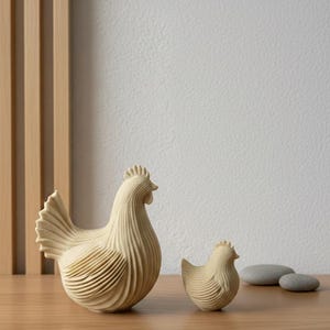 Pode incluir: Duas estatuetas de galinha na cor creme com uma superfície estriada e texturizada. A galinha maior está à esquerda, enquanto uma estatueta menor está posicionada à sua direita. Ambas as figuras apresentam um design estilizado.