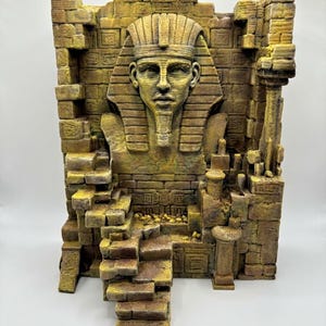 Pode incluir: Uma escultura tridimensional representando o busto de um antigo faraó egípcio centrado contra um fundo de parede de tijolos desgastados. A estrutura apresenta uma escadaria de pedra que leva à figura central, com tons texturizados de amarelo e castanho.