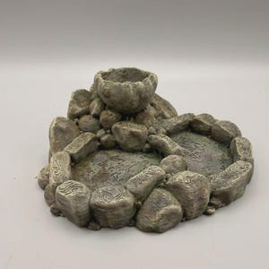 May include: A grey, textured, rock-shaped tray featuring a central elevated bowl surrounded by two lower, circular basins. The entire structure is composed of interconnected, stone-like segments with visible layered patterns and a weathered finish.