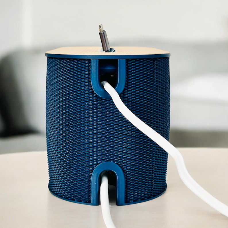 K&ouml;nnte beinhalten: Ein marineblauer Kabelorganizer mit gewebter Textur. Der zylindrische Beh&auml;lter hat eine hell holzfarbene Oberseite und ein wei&szlig;es Kabel, das von der Seite herausragt. Ein silberfarbener Stecker ist oben eingesetzt.